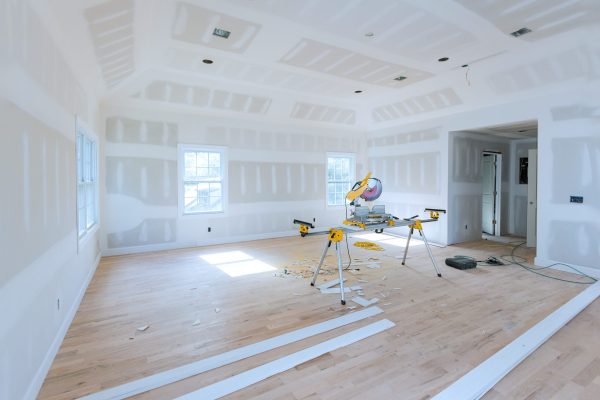 Spacious, unfinished room with drywall partially installed miter saw set up in center on stand, surrounded by wood trim pieces construction debris at hardwood floor.